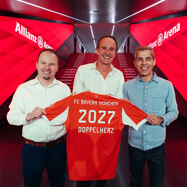 На сликата, три мажи стојат во коридор на стадионот Allianz Arena, држејќи фудбалска маичка на FC Bayern München со бројот 2027 и текстот „DOPPELHERZ“. Позадината е украсена со црвени тонови. Мажите се насмеани и изгледаат задоволно.