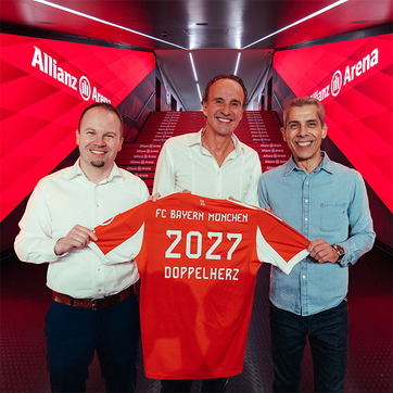 На сликата, три мажи стојат во коридор на стадионот Allianz Arena, држејќи фудбалска маичка на FC Bayern München со бројот 2027 и текстот „DOPPELHERZ“. Позадината е украсена со црвени тонови. Мажите се насмеани и изгледаат задоволно.